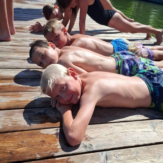 Mehrere Kinder liegen auf einem Holzsteg und genießen den sonnigen Tag. Ein Junge vorne lächelt in die Kamera.
