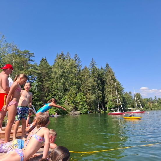 Kinder spielen am Rande eines Sees mit Booten im Wasser. Bäume säumen das Ufer und ein Junge bereitet sich auf den Sprung vor.