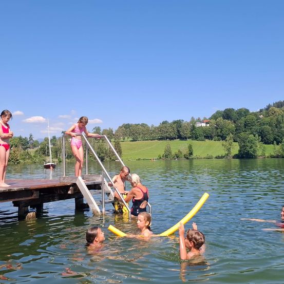 Eine Gruppe von Kindern und Erwachsenen spielt im Wasser in der Nähe einer Anlegestelle, wobei ein Mädchen in Badekleidung auf dem Steg steht.