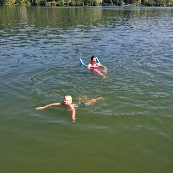 Zwei Menschen treiben in einem See. Einer trägt einen roten Schwimmring und der andere einen blauen. Bäume und ein Berg sind im Hintergrund zu sehen.