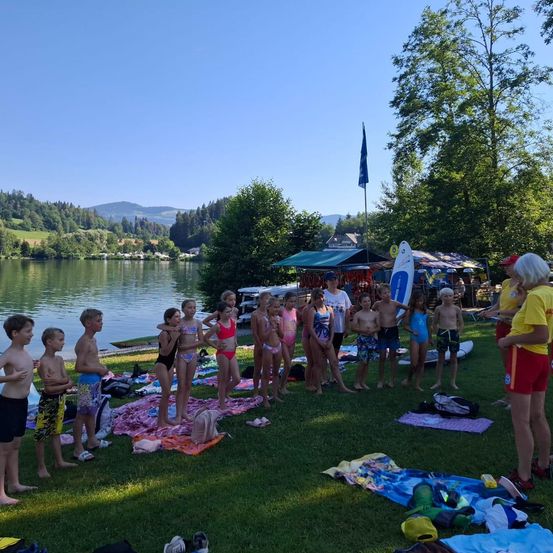 Kinder und Erwachsene in Badekleidung stehen am See, mit einer Fahne und Zelten im Hintergrund. Einige haben Handtücher und Rucksäcke auf dem Gras.