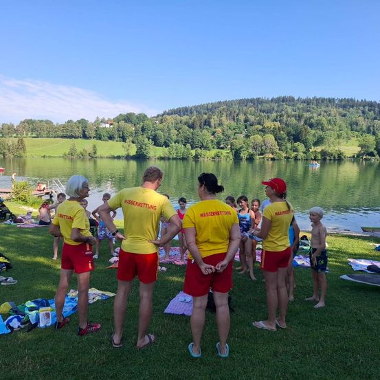 Eine Gruppe von Rettungsschwimmern steht am Ufer eines Sees, neben einer Frau und Kindern. Sie tragen gelbe Shirts mit roten Shorts. Ein Kind steht allein in ihrer Nähe.