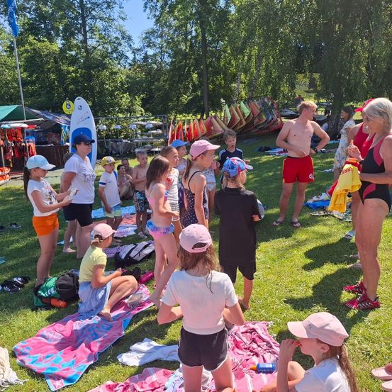 Eine Gruppe von Kindern und Erwachsenen versammelt sich auf einem Rasenfeld. Einige sitzen auf Handtüchern, während andere stehen. Surfbretter und Schwimmwesten sind im Hintergrund sichtbar. Bäume und ein Zelt befinden sich in der Ferne.