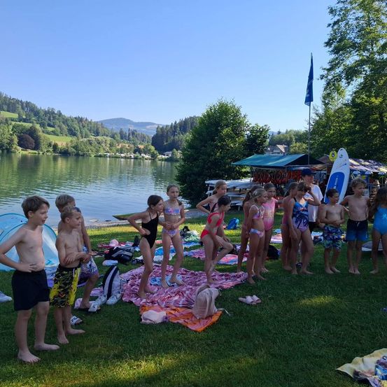 Eine Gruppe von Kindern steht am See, in Badeanzügen, mit Handtüchern und Surfbrettern auf dem Rasen. Ein Flaggenmast steht hinter einem Baldachin. Berge sind in der Ferne.