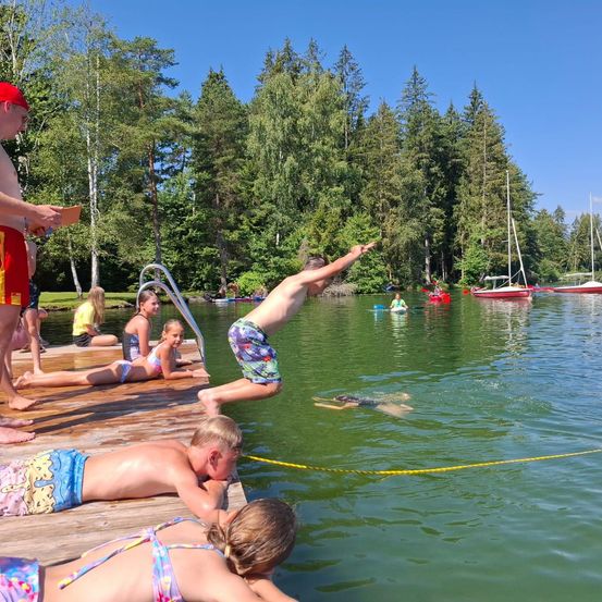 Mehrere Kinder schwimmen und spielen in einem See mit einem Steg. Einige springen ins Wasser, während andere auf dem Steg liegen. Bäume und Boote sind im Hintergrund sichtbar.