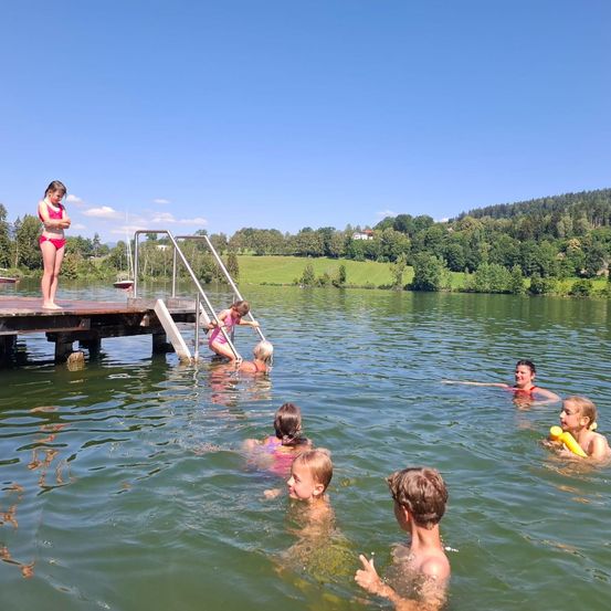 Kinder schwimmen und spielen in einem See, einige halten Schwimmhilfen. Ein Mädchen steht auf der Dock, eine Frau klettert die Treppe hoch.