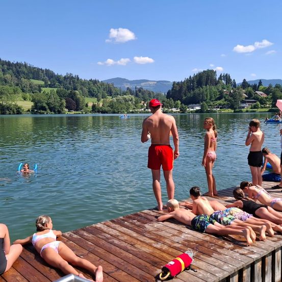 Eine Gruppe von Menschen ist auf einem Holzsteg, einige in Badeanzügen, andere im See. Ein Mann steht, während andere sich auf der Dock entspannen. Bäume und Berge sind im Hintergrund.