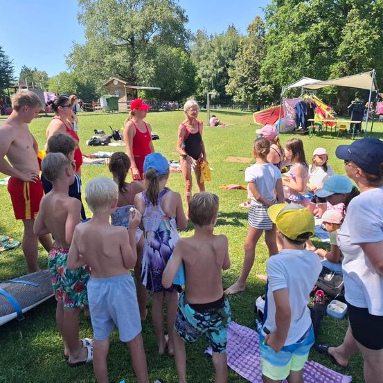 Eine Gruppe von Kindern und Erwachsenen in Badekleidung versammelt sich auf einem Rasenfeld, wahrscheinlich für einen Schwimmkurs. Einige stehen, andere sitzen auf Handtüchern. Eine Frau in einer roten Mütze gibt Anweisungen.