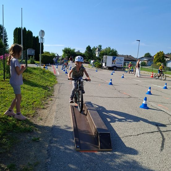 Ein junger Junge fährt ein Fahrrad eine Rampe in einer Fahrrad-Hindernisparcours hoch. In der Nähe fahren andere Kinder Fahrräder. Ein Mädchen beobachtet in der Nähe.