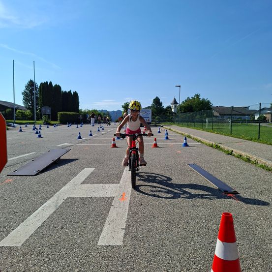 Ein Kind fährt ein Fahrrad auf einer Strecke mit mehreren Kegeln und Hindernissen in einem Außenbereich an einem sonnigen Tag.