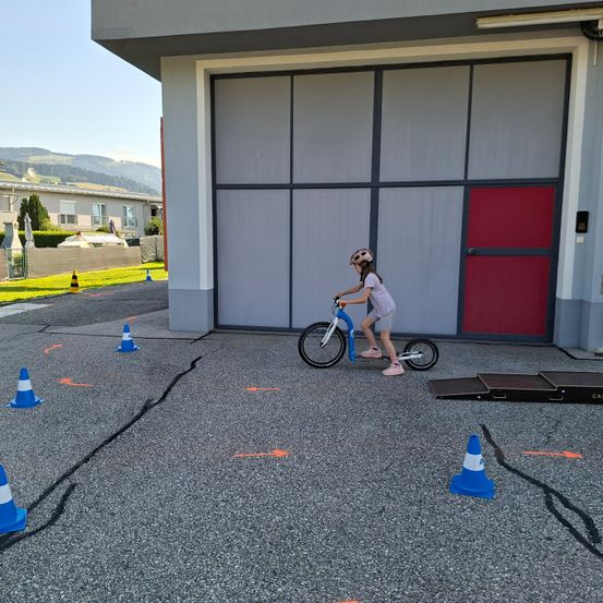 Ein Kind fährt auf einer Strecke mit Hindernissen ein Fahrrad, in der Nähe einer Garage. Blaue und orangefarbene Kegel säumen den Weg.