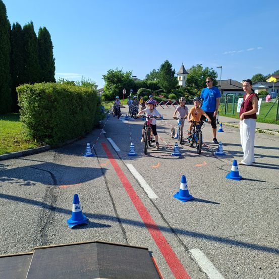 Eine Gruppe von Kindern fährt auf einer Straßenstrecke mit Fahrrädern, begleitet von Erwachsenen. Verkehrskegel markieren den Kurs, und es gibt Bäume und einen Zaun im Hintergrund.