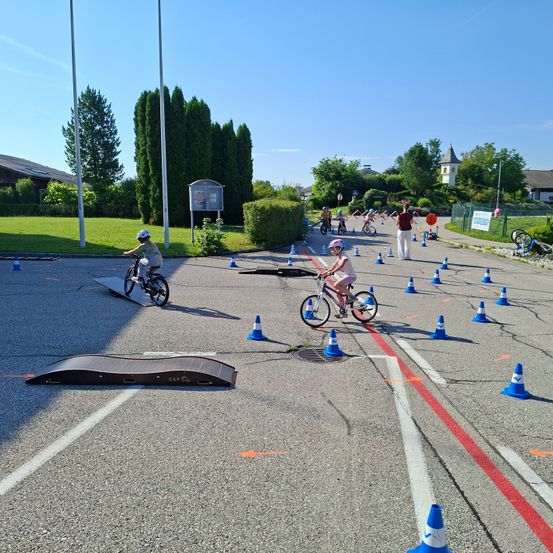 Kinder fahren mit Fahrrädern durch einen Parcours mit blauen und orangefarbenen Kegeln. Zwei Kinder fahren auf einer Rampe. Eine Person in Rot steht am Rand. Bäume und Gebäude sind im Hintergrund.