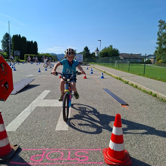 Ein Kind mit Helm fährt ein Fahrrad auf einer asphaltierten Straße mit Verkehrskegel und Markierungen. Dahinter sind andere Radfahrer in der Ferne. Bäume, Häuser und ein Turm sind im Hintergrund sichtbar.