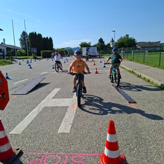 Eine Gruppe von Kindern fährt auf einer Straße mit Fahrrädern, umgeben von Verkehrskegeln. Ein Kind trägt einen blauen Helm und ein orangefarbenes Shirt.