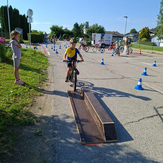 Ein Junge fährt ein Fahrrad über eine Rampe, umgeben von anderen Kindern und Hütchen. Ein Mädchen beobachtet ihn vom Gras.