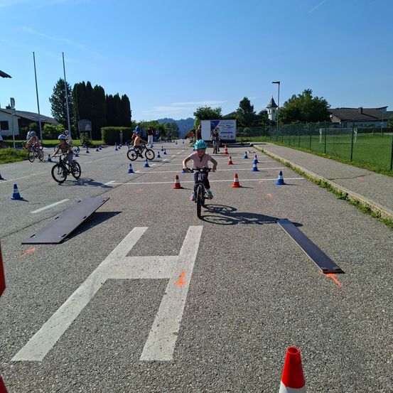 Eine Gruppe von Kindern fährt mit Fahrrädern durch einen Parcours mit orangefarbenen Kegel. Der Parcours ist mit einem H auf der Straße markiert. Bäume und Gebäude sind im Hintergrund.