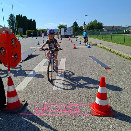 Ein kleines Kind fährt auf einem Kurs mit Verkehrskegeln ein Fahrrad. Im Hintergrund sind andere Kinder auf Fahrrädern. Das Wort 'STOP' ist auf dem Boden gemalt.
