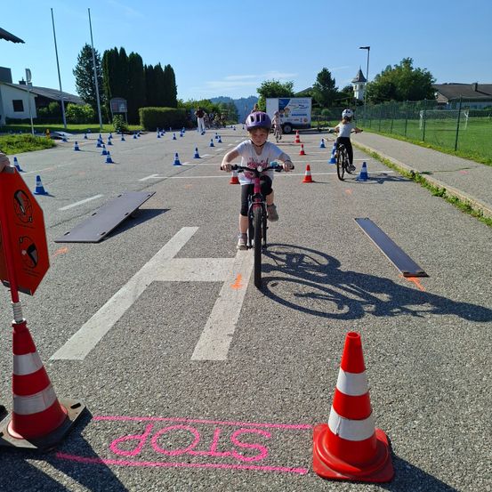 Kinder fahren auf einer markierten Straße mit Fahrrädern, umgeben von Verkehrskegeln und einem Schild mit der Aufschrift '2015'. Bäume und ein Gebäude sind in der Ferne zu sehen.