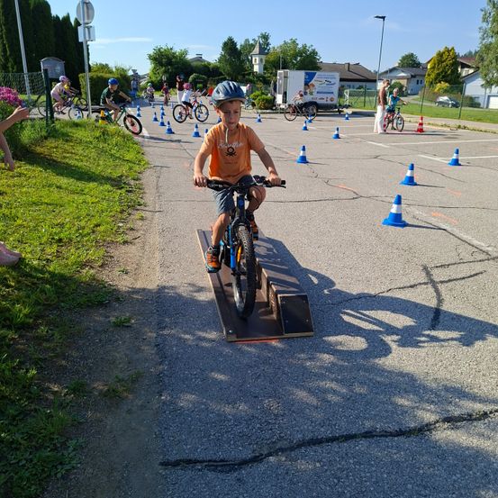 Ein Junge im Helm fährt ein Fahrrad auf einer Rampe über Hütchen auf einer asphaltierten Fläche. In der Nähe fahren mehrere andere Kinder mit Fahrrädern.