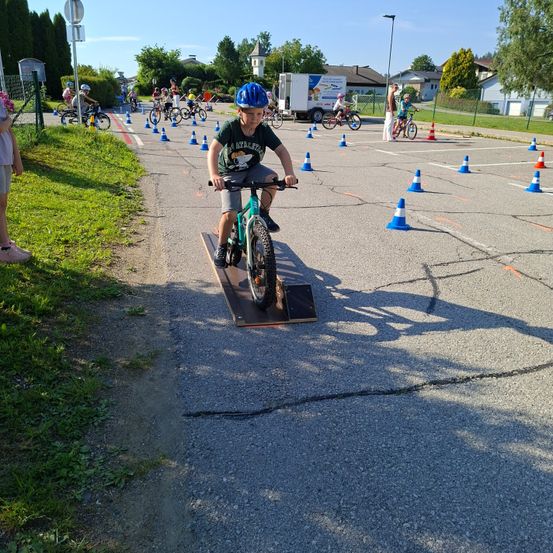 Ein Junge fährt ein Fahrrad über eine Rampe in einem Parcours. Verkehrskegel umgeben den Kurs. Viele Kinder fahren in der Nähe Fahrräder.