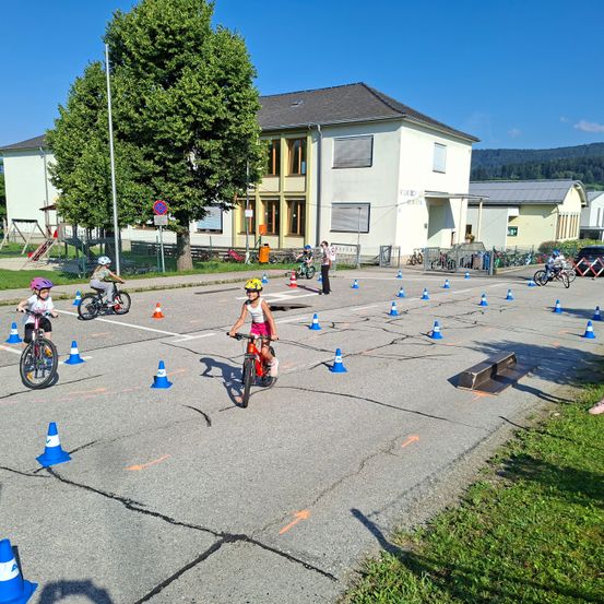 Kinder fahren mit Fahrrädern in einem Parkplatz, wo Kegel in einem Zickzack-Muster angeordnet sind. Dahinter sind ein Gebäude und ein Spielplatz zu sehen.
