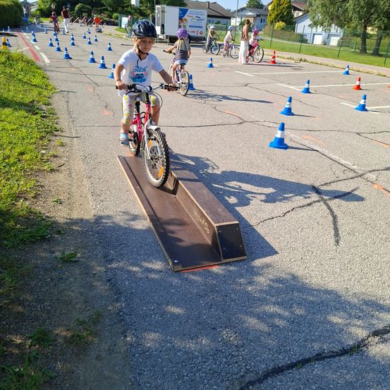 Ein Kind fährt ein Fahrrad auf einer Rampe in einem Hindernisparcours. Andere Kinder navigieren durch Kegel auf der Straße.