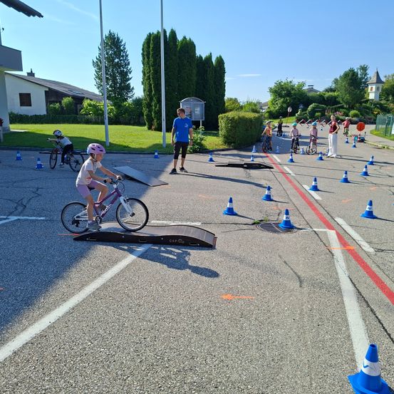 Eine Gruppe von Kindern fährt mit Fahrrädern auf einem Parkplatz mit Verkehrskegeln. Ein Mädchen ist auf einer Rampe. Ein Mann steht in der Nähe, und eine Frau beobachtet in der Ferne.