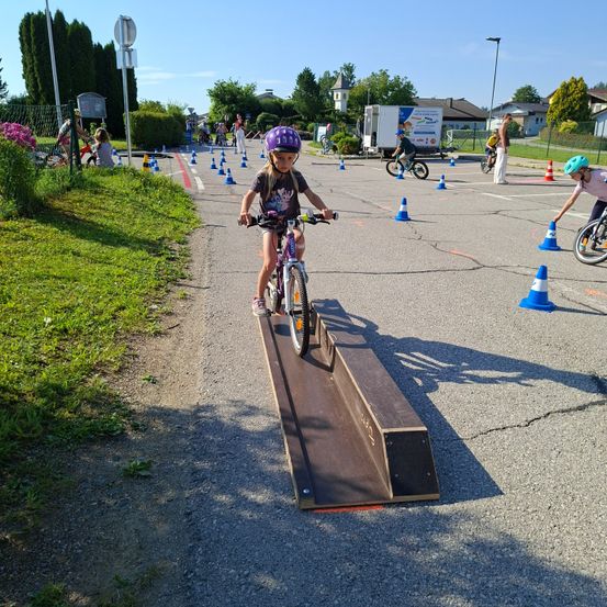 Ein kleines Mädchen in einem lila Helm fährt mit einem Fahrrad über eine Rampe mit blauen und weißen Kegeln an den Seiten.