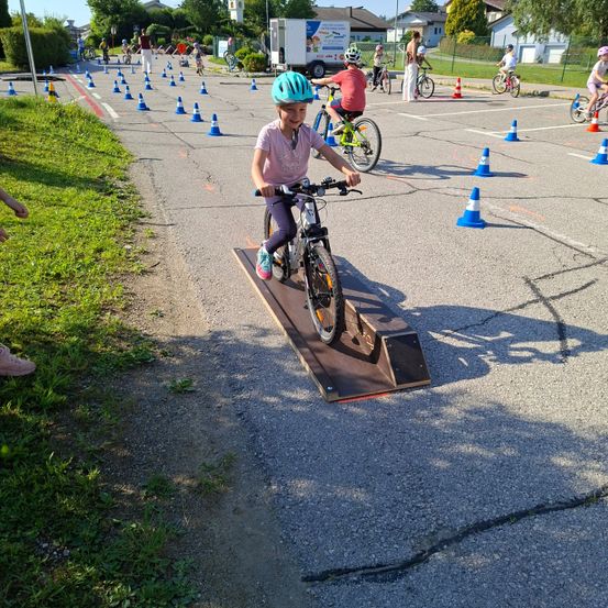 Ein Kind in einem Helm fährt ein Fahrrad über eine Rampe, umgeben von Verkehrskegeln. Andere Kinder fahren Fahrräder auf einem Parkplatz.