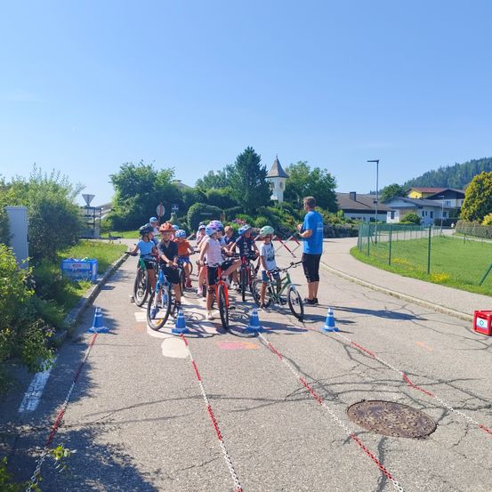 Eine Gruppe von Kindern fährt in einer Reihe auf einem asphaltierten Weg auf Fahrrädern, begleitet von einem Mann in Blau. Kegel markieren den Weg. Bäume und Gebäude sind im Hintergrund zu sehen.