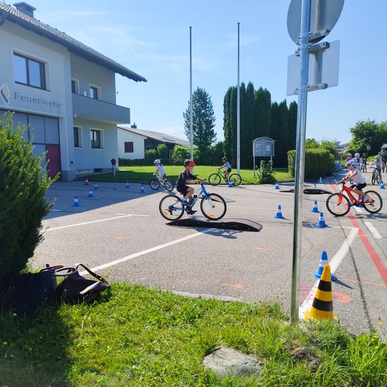 Mehrere Kinder fahren in einer Reihe mit orangefarbenen und blauen Kegeln und einer Rampe im Parkplatz auf Fahrrädern. Ein Gebäude mit der Aufschrift Feuerwehr steht links.