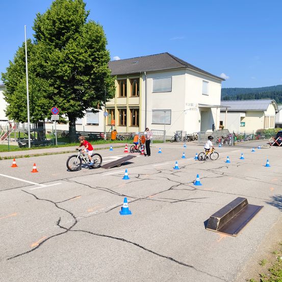 Kinder fahren Fahrräder durch einen Parcours aus Hütchen in einem offenen Bereich mit einem Gebäude im Hintergrund.