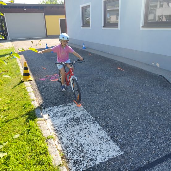 Ein junges Kind mit Helm fährt ein rotes Fahrrad auf einem gepflasterten Weg. Ein Gebäude und Verkehrskegel sind in der Nähe.