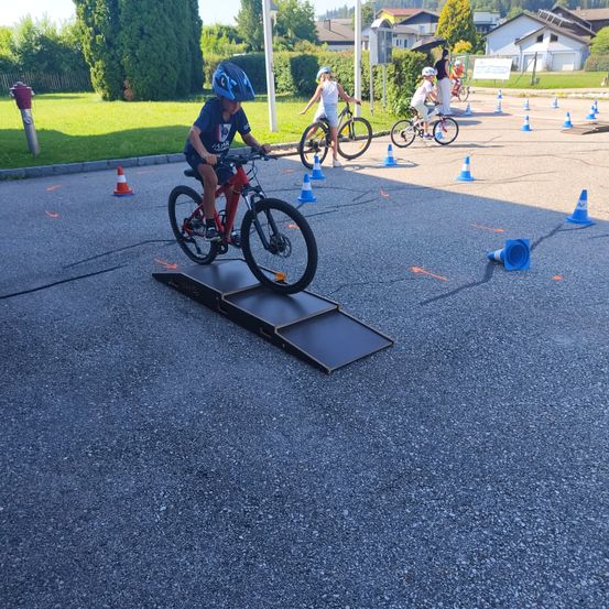 Mehrere Kinder fahren auf einem gepflasterten Bereich mit einer Rampe Fahrrad, umgeben von Verkehrskegel. Ein Kind fährt auf der Rampe.