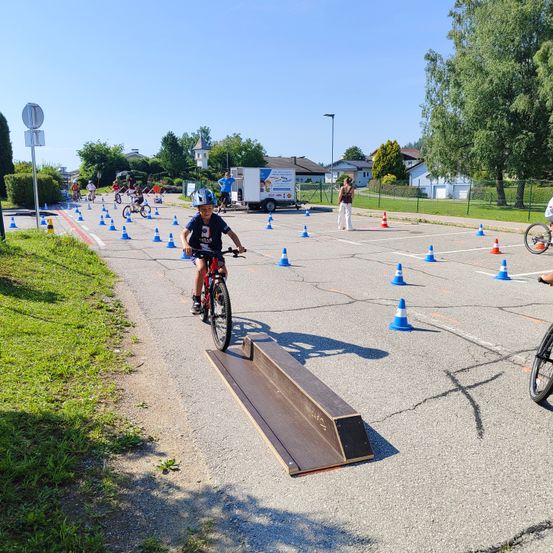 Eine Gruppe von Kindern fährt auf einer mit Kegeln markierten Straße mit Fahrrädern. Ein Junge fährt auf einer Rampe.