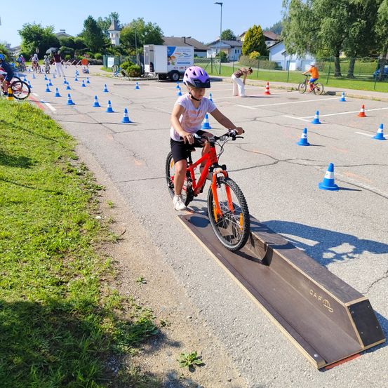 Ein Kind fährt ein rotes Fahrrad über eine Rampe mit orangefarbenen Hütchen auf einer Straße. Ein weißer LKW ist in der Nähe geparkt.