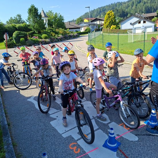 Eine Gruppe von Kindern in Helmen steht in einer Reihe auf der Straße und fährt mit Fahrrädern, ein Mann gibt vorne Anweisungen.
