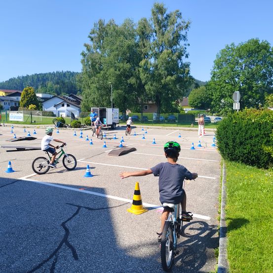 Kinder fahren auf einem gepflasterten Bereich mit vielen blauen Hütchen und Hindernissen mit Fahrrädern. Ein Junge mit Helm bereitet sich auf das Wenden seines Fahrrads vor.