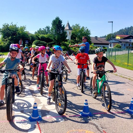 Eine Gruppe von Kindern fährt mit Fahrrädern auf einer Straße mit Kegel. Sie tragen Helme und haben einen Mann, der sie leitet. Bäume und Gebäude sind im Hintergrund.