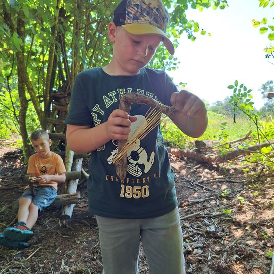 Ein Junge steht in einem Wald und hält ein handgefertigtes Werkzeug, möglicherweise einen Bogen. Ein anderer Junge sitzt auf einem Baumstamm hinter ihm. Das Gebiet ist von Bäumen und trockenen Blättern umgeben.