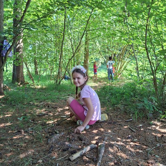 Ein kleines Mädchen sitzt im Wald und lächelt für ein Foto. Dahinter gehen zwei Kinder in der Ferne. Bäume und Grün umgeben sie.