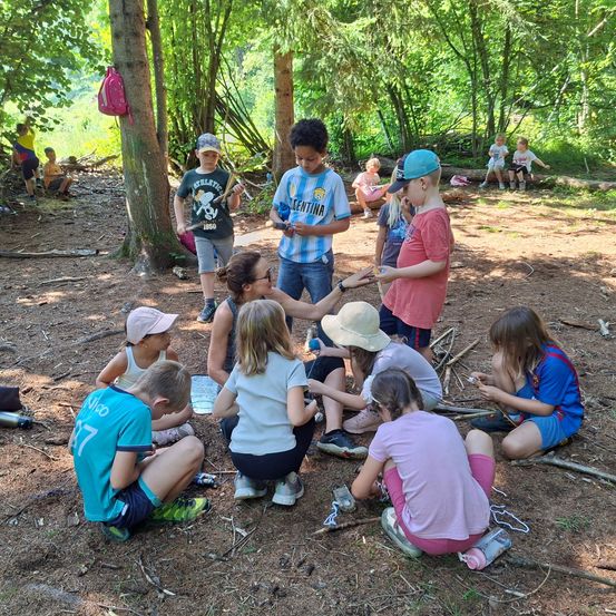 Kinder und Erwachsene versammeln sich in einem Waldgebiet, einige sitzen auf dem Boden. Eine Frau steht und spricht mit einer Gruppe von Kindern. Es gibt Bäume und Pflanzen im Bereich.