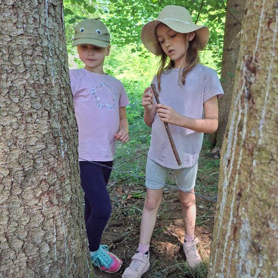 Zwei Mädchen erkunden einen Waldweg, eine steht neben einem Baum und die andere hält einen Stock. Sie tragen Hüte und Turnschuhe und genießen die Natur.