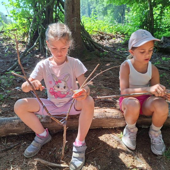 Zwei Mädchen basteln mit Stöcken auf einem Baumstamm im Wald. Ein Mädchen konzentriert sich auf ihr Projekt, während das andere zuschaut.