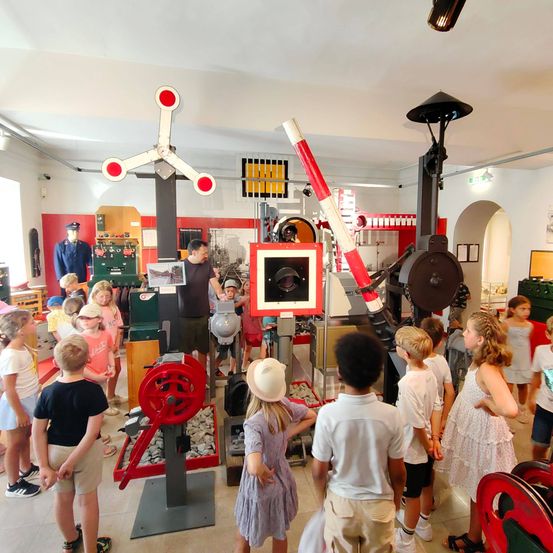 Eine Gruppe von Kindern erkundet eine Ausstellung in einem Museum. Sie stehen um Maschinen und Geräte herum, ein Mann unterrichtet sie.