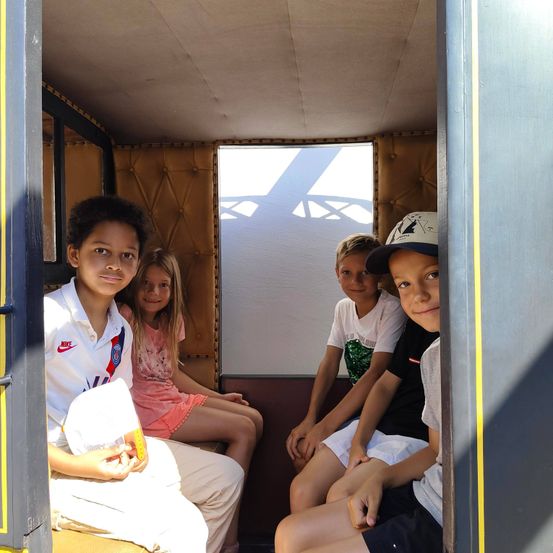 Vier Kinder sitzen in einem Wagen. Ein Junge hält einen weißen Hut. Ein Mädchen in einem rosa Kleid sitzt neben ihm. Zwei Jungen sitzen ihnen gegenüber. Sonnenlicht scheint durch ein Fenster.