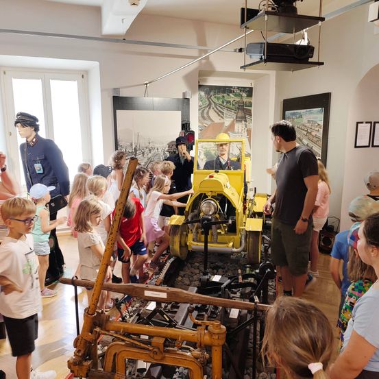 Eine Gruppe von Kindern und Erwachsenen versammelt sich in einem Museum um ein gelbes Zugmodell, ein Mann erklärt ihnen etwas.
