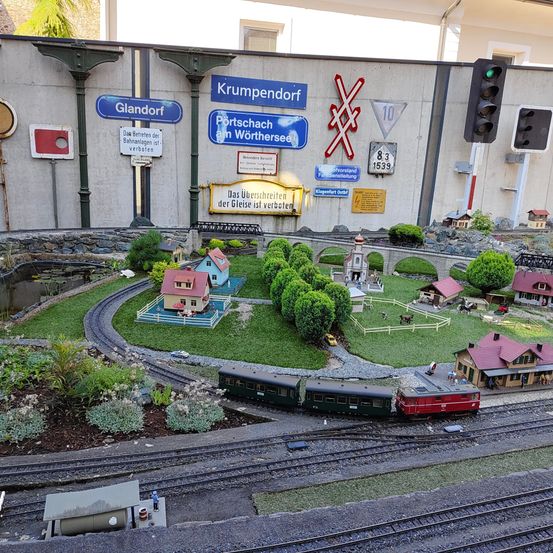 Eine Miniatur-Modelleisenbahn mit einem Bahnhof, Häusern und einer kleinen Brücke. Sie befindet sich auf einer Strecke mit einem rot-schwarzen Zug, der sich darauf bewegt. Die Wand dahinter hat mehrere Schilder.