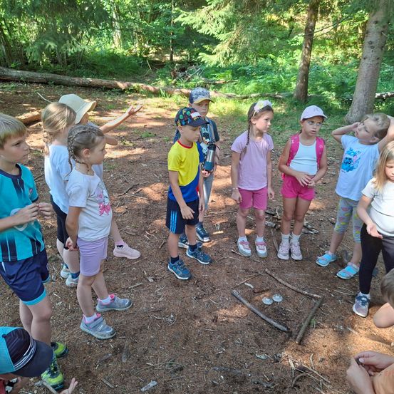 Eine Gruppe von Kindern steht in einem Wald, einige mit Hüten und Turnschuhen, schauen neugierig herum. Ein kleiner Kreis aus Ästen und Steinen liegt auf dem Boden.